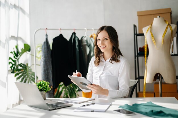 Mujer diseñadora de moda hablando con un teléfono inteligente y usando una computadora portátil con una tableta digital en un estudio moderno la ropa colgando en los estantes a la luz de la mañana