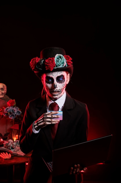 Mujer disfrazada de dama de la muerte comprando en línea con tarjeta de crédito y laptop, usando traje y sombrero con maquillaje de calavera. Pareciendo diosa de la muerte y santa muerte, dios de los muertos.