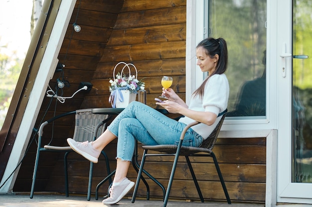 La mujer disfruta del clima cálido sentada en la terraza de la cabaña de madera con un teléfono inteligente en la naturaleza