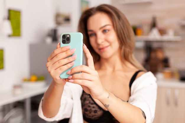 Mujer disfrutando tomando fotos durante el desayuno con ropa interior sexy. Mujer seductora con tatuajes con smartphone usando lencería temping en la mañana.