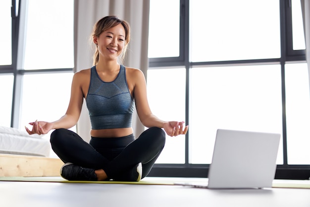 Mujer disfrutando de yoga en la estera sola en casa, dando un taller de yoga educativo en línea, usando una computadora portátil. mujer fuerte en ropa deportiva