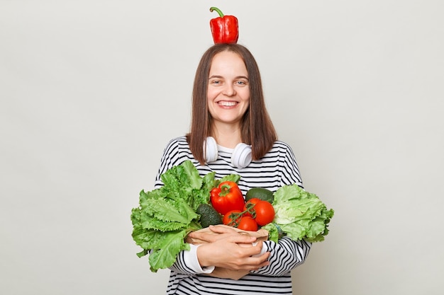 Una mujer divertida y alegre con el pelo castaño y un pantalones a rayas que se encuentra aislada sobre un fondo gris sosteniendo verduras posando con pimienta en la cabeza mirando la cámara