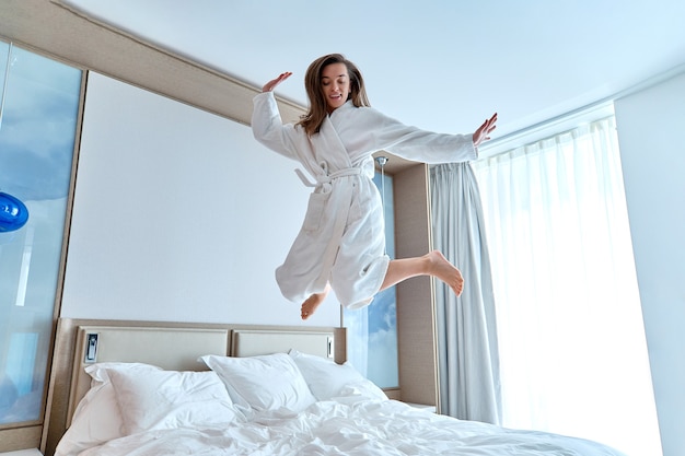 Mujer divertida alegre satisfecha en bata de baño saltando sobre la cama en una habitación de hotel durante un momento feliz. Sentirse bien y disfrutar del concepto de vida. Estilo de vida fácil