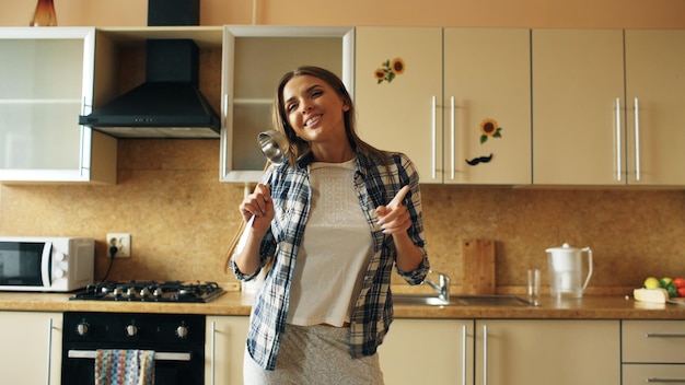 mujer divertida bailando y cantando con cucharón mientras tiene tiempo libre en la cocina en casa