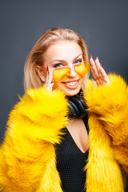 Mujer divertida feliz con auriculares en gafas de sol amarillas en abrigo de piel amarillo de moda en gris