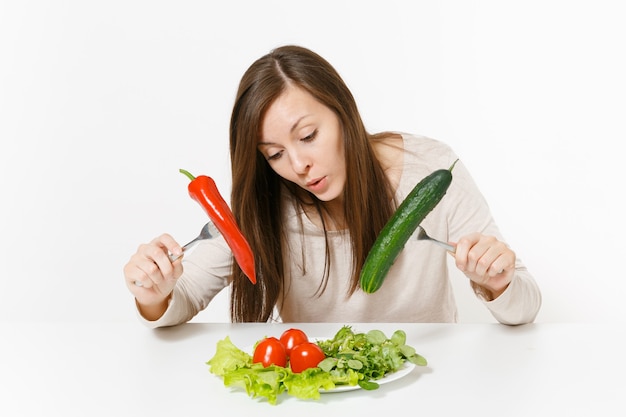 Foto mujer divertida en la mesa con ensalada de hojas de lechuga en un plato, pimiento, pepino en horquillas aisladas sobre fondo blanco. nutrición adecuada, comida vegetariana, concepto de dieta de estilo de vida saludable. área para copiar espacio.