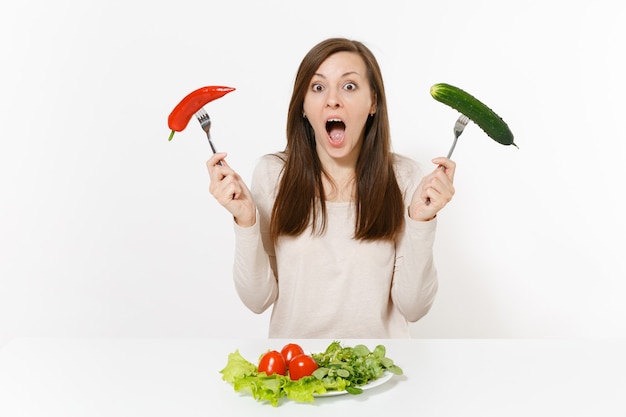 Mujer divertida en la mesa con ensalada de hojas de lechuga en un plato, pimiento, pepino en horquillas aisladas sobre fondo blanco. Nutrición adecuada, comida vegetariana, concepto de dieta de estilo de vida saludable. Área para copiar espacio.