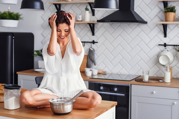 Una mujer divertida con el pelo rizado y vestida con una bata blanca siente pánico en la cocina mientras cocina.