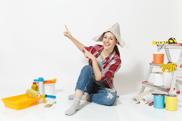 Mujer divertida con sombrero de periódico apuntando con el dedo índice hacia arriba en el espacio de la copia, sentarse en el piso con instrumentos para el apartamento de renovación aislado sobre fondo blanco. Papel tapiz, herramientas de pintura. Reparar el concepto de hogar.