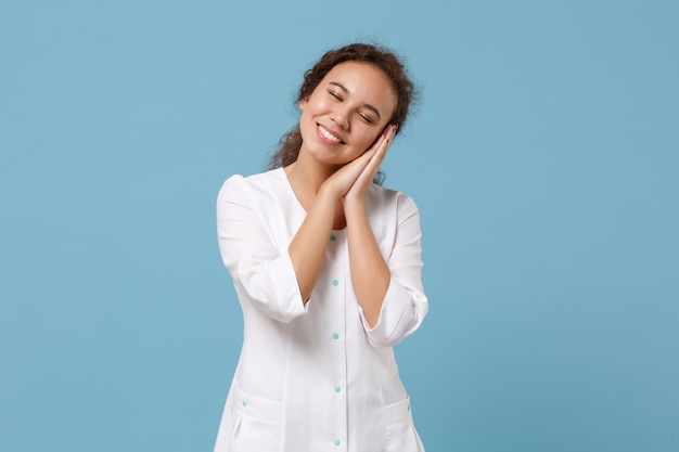 Mujer doctora afroamericana aislada de fondo azul. Una doctora con bata médica blanca duerme con las manos dobladas debajo de la mejilla. Concepto de medicina sanitaria del personal sanitario. Simulacros de espacio de copia.