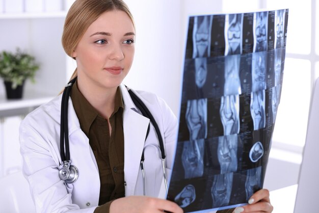 Mujer doctora examinando una imagen de rayos X cerca de la ventana en el hospital. Cirujano u ortopedista en el trabajo. Concepto de medicina y salud. La blusa de color caqui de un terapeuta se ve bien.