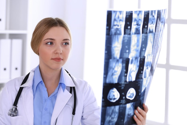 Mujer doctora examinando una imagen de rayos X cerca de la ventana en el hospital. Cirujano u ortopedista en el trabajo. Concepto de medicina y salud. Se ve bien la blusa de color azul de un terapeuta.