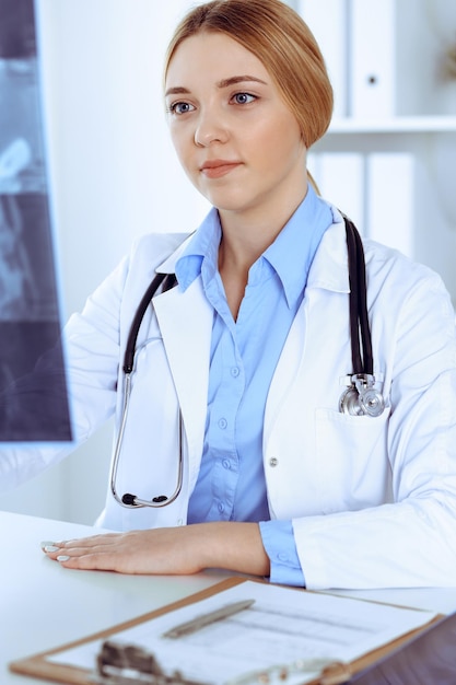 Mujer doctora examinando una imagen de rayos X cerca de la ventana en el hospital. Cirujano u ortopedista en el trabajo. Concepto de medicina y salud. Se ve bien la blusa de color azul de un terapeuta.