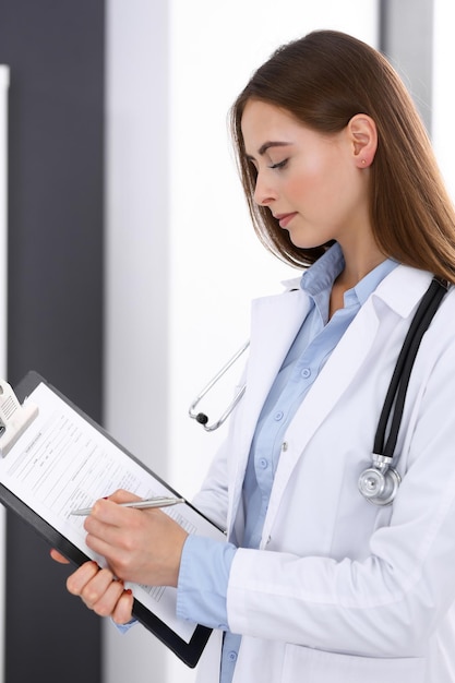 Mujer doctora llenando un formulario médico mientras está de pie cerca de la ventana en la oficina del hospital. Médico feliz en el trabajo. Concepto de medicina y atención de la salud.