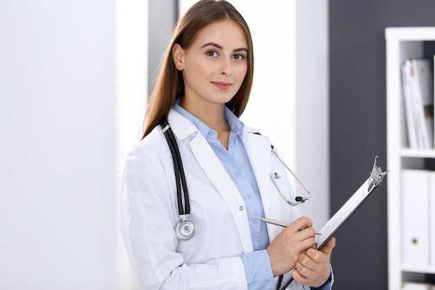 Mujer doctora llenando un formulario médico mientras está de pie cerca de la ventana en la oficina del hospital. Médico feliz en el trabajo. Concepto de medicina y atención de la salud.