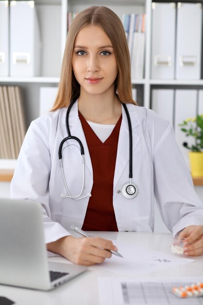 Mujer doctora llenando un formulario médico o escribiendo en una laptop mientras está sentada en la mesa cerca de la ventana. Concepto de medicina y atención de la salud.