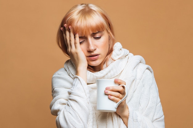 Mujer con dolor de cabeza, sosteniendo una taza de bebida caliente, tocando su frente, sintiendo los primeros síntomas de gripe