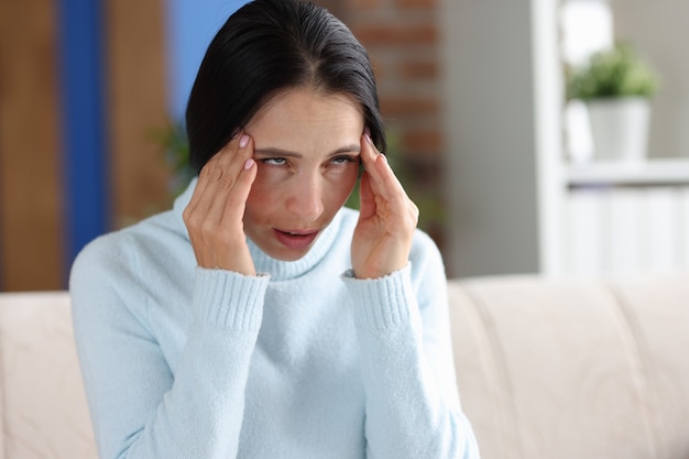 Mujer con dolor de cabeza tiene las manos en las sienes mientras está sentado en el sofá. Síntomas de migraña en la cabeza