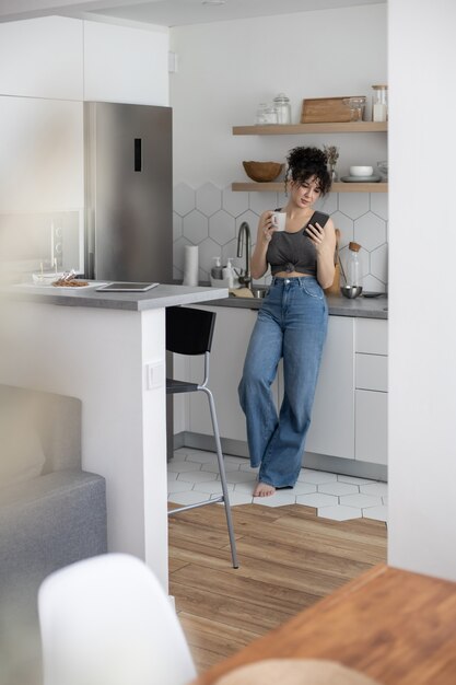 Foto mujer doméstica agradable que se relaja en la cocina moderna charlando utiliza el interior de la cocina blanca del smartphone