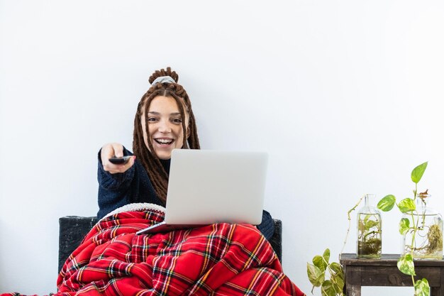 Mujer con dreadlocks trabajando con una computadora portátil en casa y con un control remoto en la mano