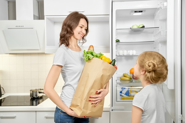 Mujer e hija con bolsa de productos cerca de nevera vacía.