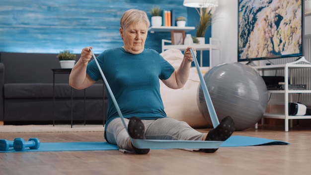 Mujer de edad con banda de resistencia para entrenar los músculos en la estera de yoga. Persona jubilada haciendo ejercicio físico y tirando de cinturón elástico, entrenando con equipos para entrenamiento físico. Pensionista
