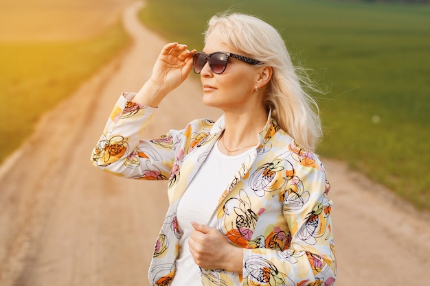 Mujer de edad con una chaqueta en la carretera en el campo verde. Sostiene gafas de sol.