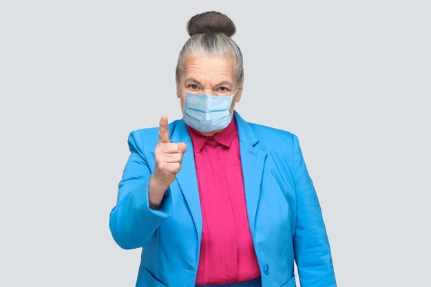 Mujer de edad enojada con máscara médica quirúrgica advirtiéndole. Abuela con traje celeste y camisa rosa de pie con el pelo gris con moño recogido. Foto de estudio de interior, aislado sobre fondo gris