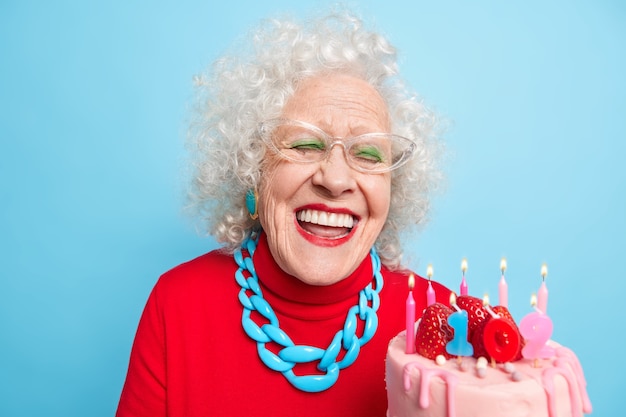 Mujer de edad positiva con cabello gris y rizado sonríe ampliamente tiene dientes blancos perfectos sostiene pastel celebra cumpleaños tiene edad de jubilación