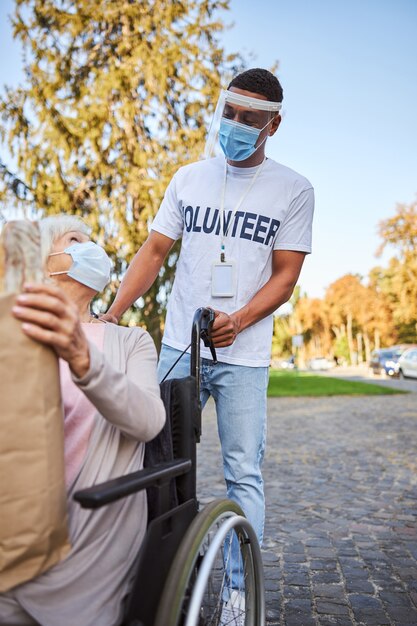 Mujer de edad en una silla de ruedas con una máscara médica que se dirige a un voluntario en un escudo protector y sosteniendo una bolsa de compras