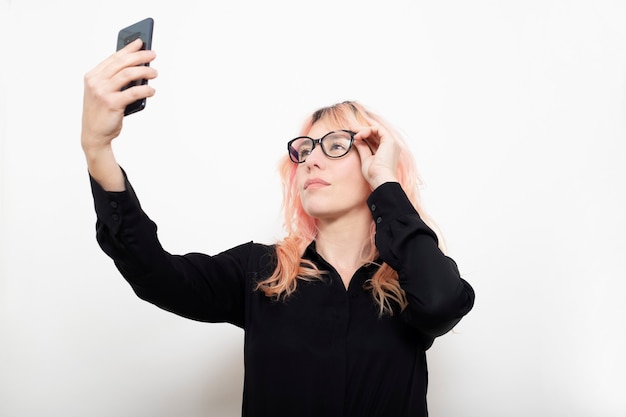 Mujer ejecutiva ajustando sus gafas con el espejo de su teléfono móvil sobre fondo blanco.