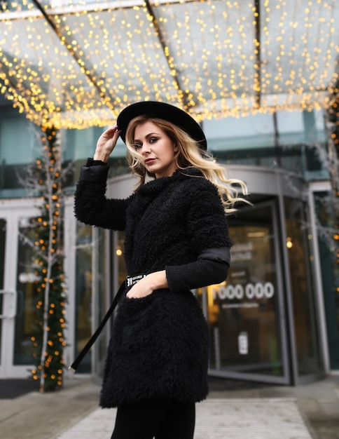 Mujer elegante en un abrigo negro cálido y un sombrero en la calle con un fondo de luz navideña