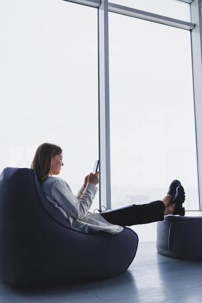 Mujer elegante analizando gráficos en aparatos y concentrándose en la pantalla del teléfono mientras se sienta junto a una ventana de pared completa en una oficina moderna