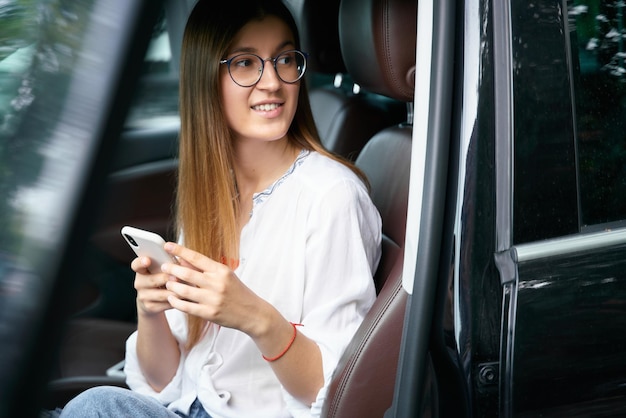 Mujer elegante con anteojos sentada en el auto y mirando hacia otro lado usando un smartphone. Transportación