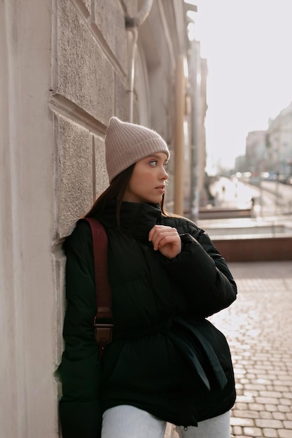 Una mujer elegante y atractiva con gorra y chaqueta oscura está parada en una calle soleada en el centro de la ciudad y mirando a un lado