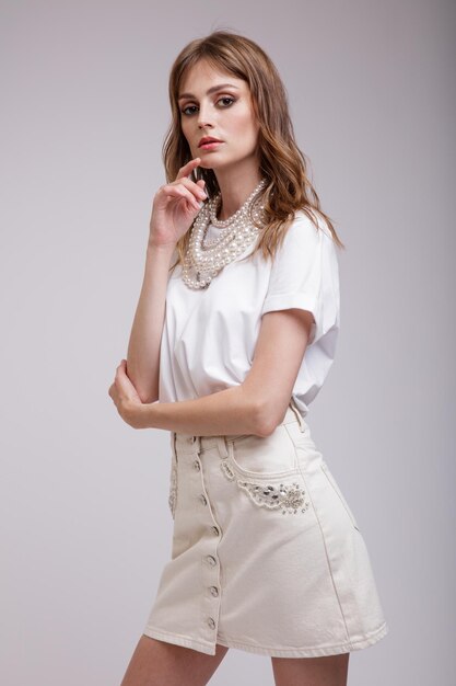 Mujer elegante en bastante collar de perlas de camiseta de falda de mezclilla blanca posando sobre fondo gris suave