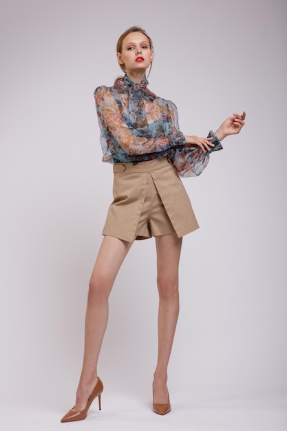 Mujer elegante en blusa azul con lazo estampado de flores en el cuello shorts beige sobre fondo blanco.