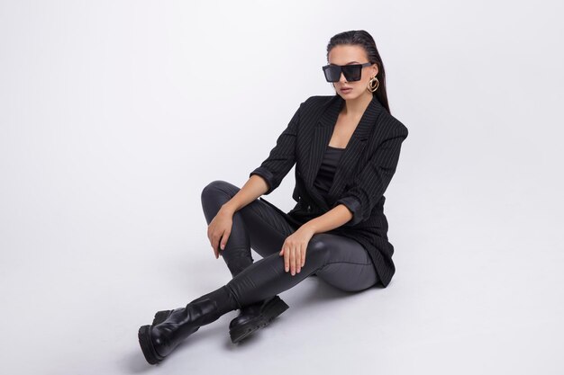 mujer elegante con bonita chaqueta negra, pantalones de cuero, botas, gafas de sol sobre fondo blanco.