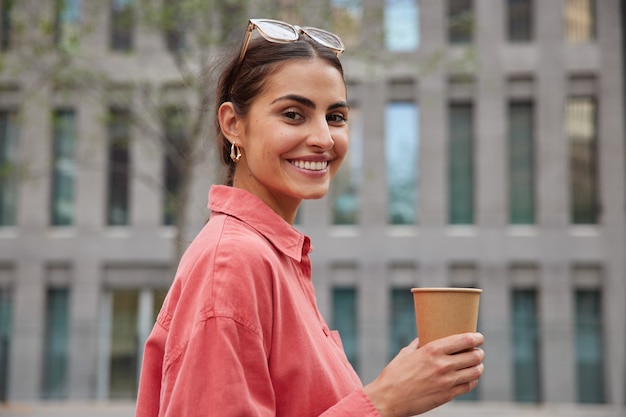 Mujer con elegante camisa roja, gafas de sol en la cabeza, paseos en una ciudad desconocida, bebe café de una taza de papel, disfruta del tiempo libre, sonríe, plantea ampliamente contra el edificio borroso