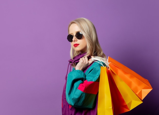 Mujer elegante en cazadora de los años 80 y gafas de sol roud sostiene bolsas de compras en la pared púrpura