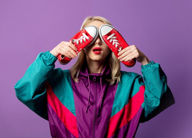 Mujer elegante en cazadora de los años 80 y gafas de sol roud sostiene gumshoes rojos en la pared púrpura