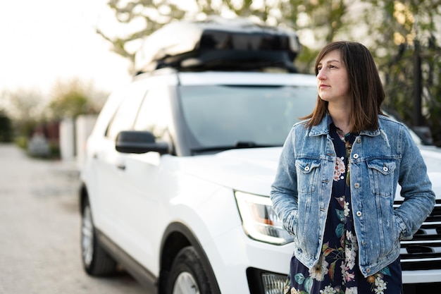 Mujer elegante con chaqueta de jeans de pie cerca de un auto todoterreno americano blanco con caja portaequipajes en el techo