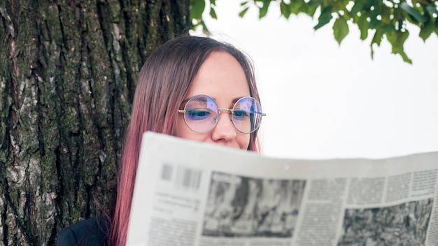 Mujer elegante concentrada leyendo el periódico en el parque Empresaria joven enfocada con cabello largo teñido en ropa formal y anteojos de pie cerca de un árbol y leyendo el periódico con interés