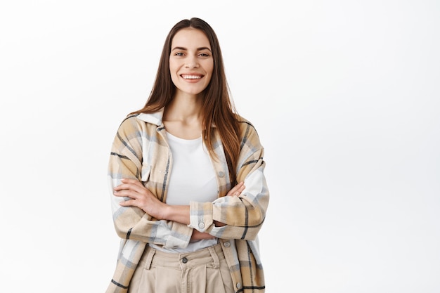 Mujer elegante cruza los brazos en el pecho y sonríe, se ve segura y asertiva, de pie como una pared blanca profesional