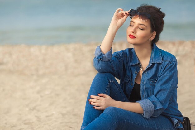 Una mujer elegante disfruta del sol sosteniendo gafas de sol y una pequeña bolsa cruzada de cuero sentada en la playa