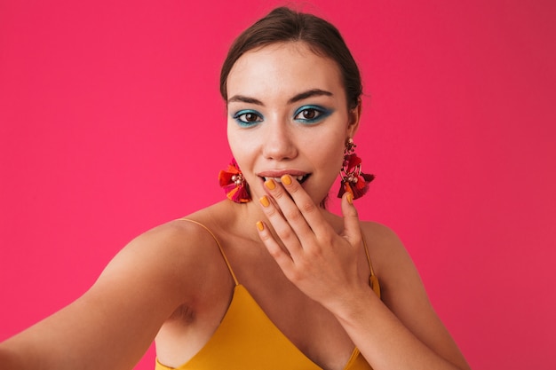 Mujer elegante emocionada con aretes que cubren la boca con la mano mientras toma selfie