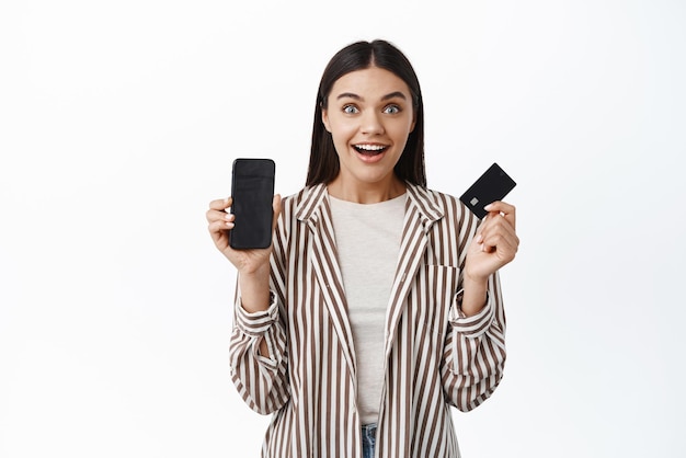 Mujer elegante emocionada que muestra la pantalla vacía del teléfono inteligente y la tarjeta de crédito de plástico que se ve feliz y sonriente publicidad fondo blanco de la tienda en línea