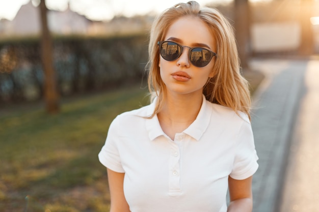 Mujer elegante hermosa joven inconformista en gafas de sol de moda en una camiseta polo blanca está de pie en la calle de un atardecer de verano. Rubia linda chica de moda.