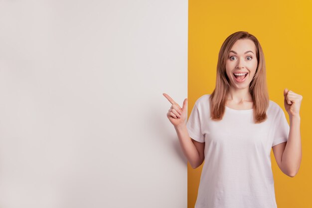 Mujer elegante joven que señala el tablero blanco del espacio vacío levantar el puño en la pared amarilla