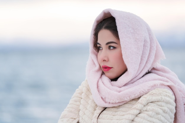 Mujer elegante con labios carnosos rojos en abrigo de piel blanco y bufanda rosa en la cabeza mirando hacia otro lado